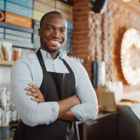 Handsome Black African American Barista with Short Hair and Beard Wearing Apron is Smiling in Coffee Shop Restaurant. Portrait of Happy Employee Behind Cozy Loft-Style Cafe Counter.
