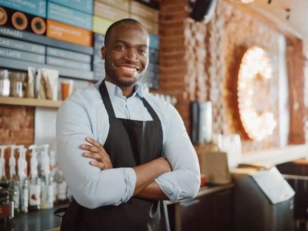 Handsome Black African American Barista with Short Hair and Beard Wearing Apron is Smiling in Coffee Shop Restaurant. Portrait of Happy Employee Behind Cozy Loft-Style Cafe Counter.