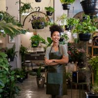 Portrait of african american woman with crossed arms wearing apron standing in botanical store. Smiling young woman in botany store standing between plants looking at camera. Happy small business owner working at flower shop standing surrounded by plants.